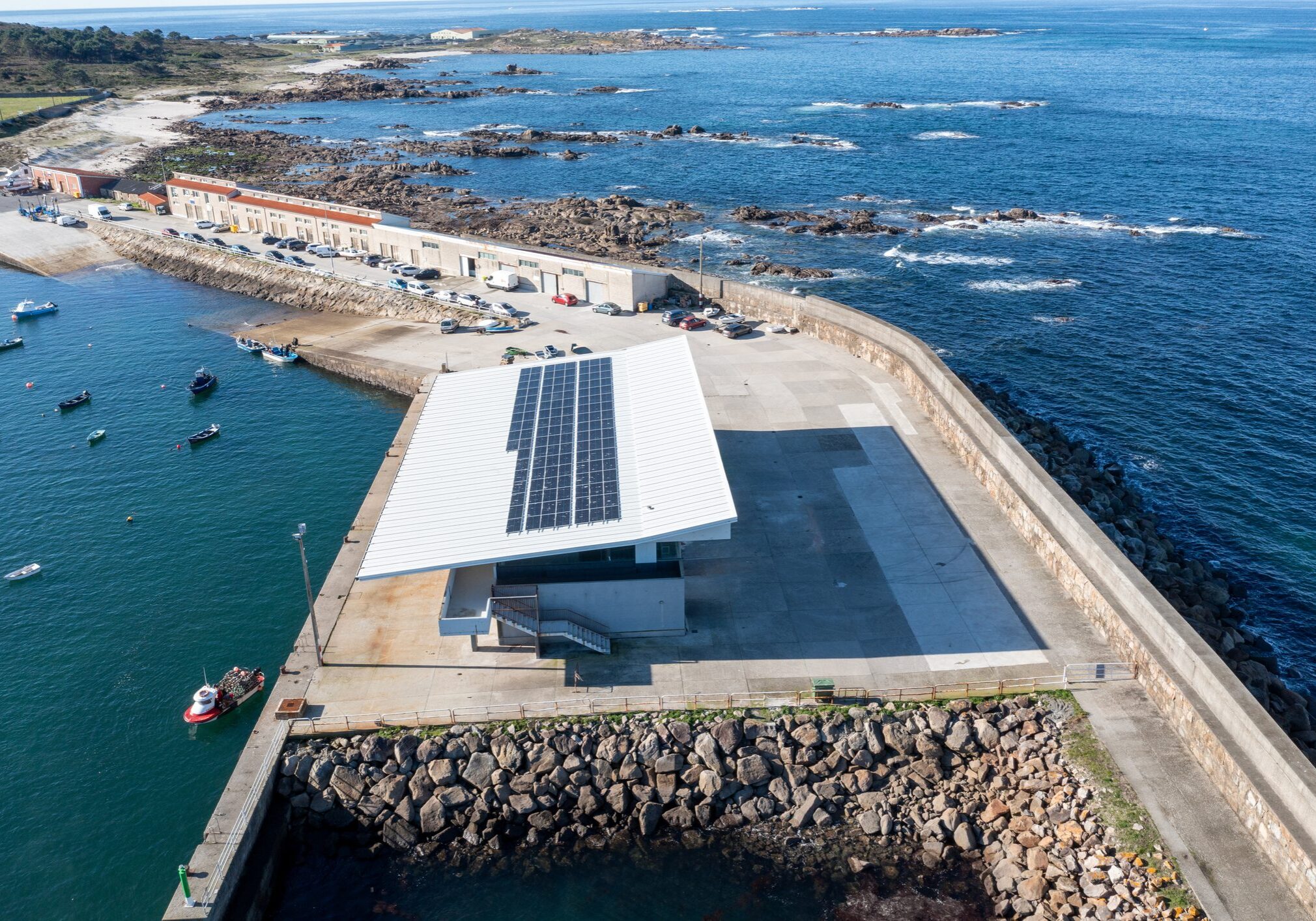 Aerial view of a modern building with solar panels on its roof, located at the edge of a pier in a harbor with boats and rocky coastline